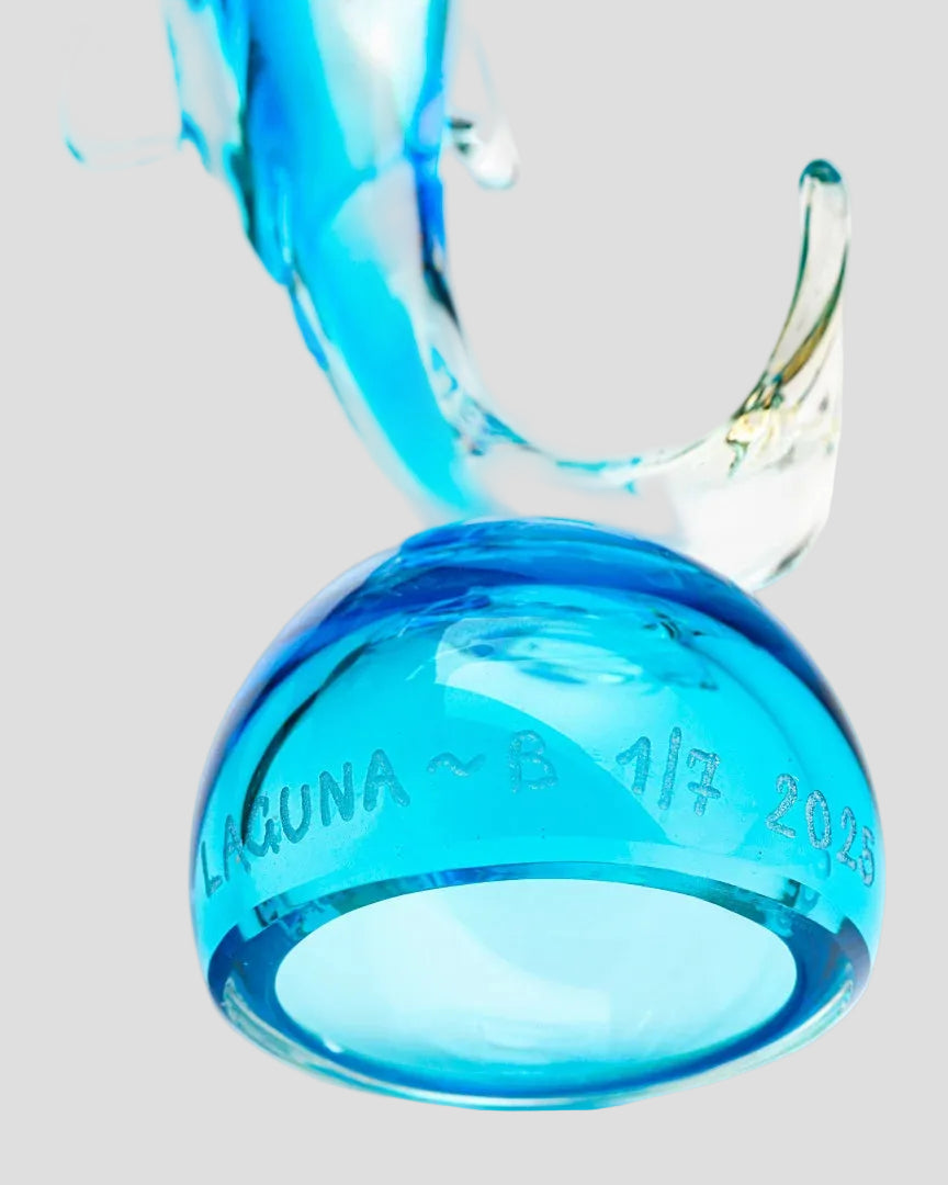 A translucent blue glass ring-shaped object with etched text reading 'LAGUNA ~ 8 1/7 2013' on a white background, with a curved clear glass element behind it.