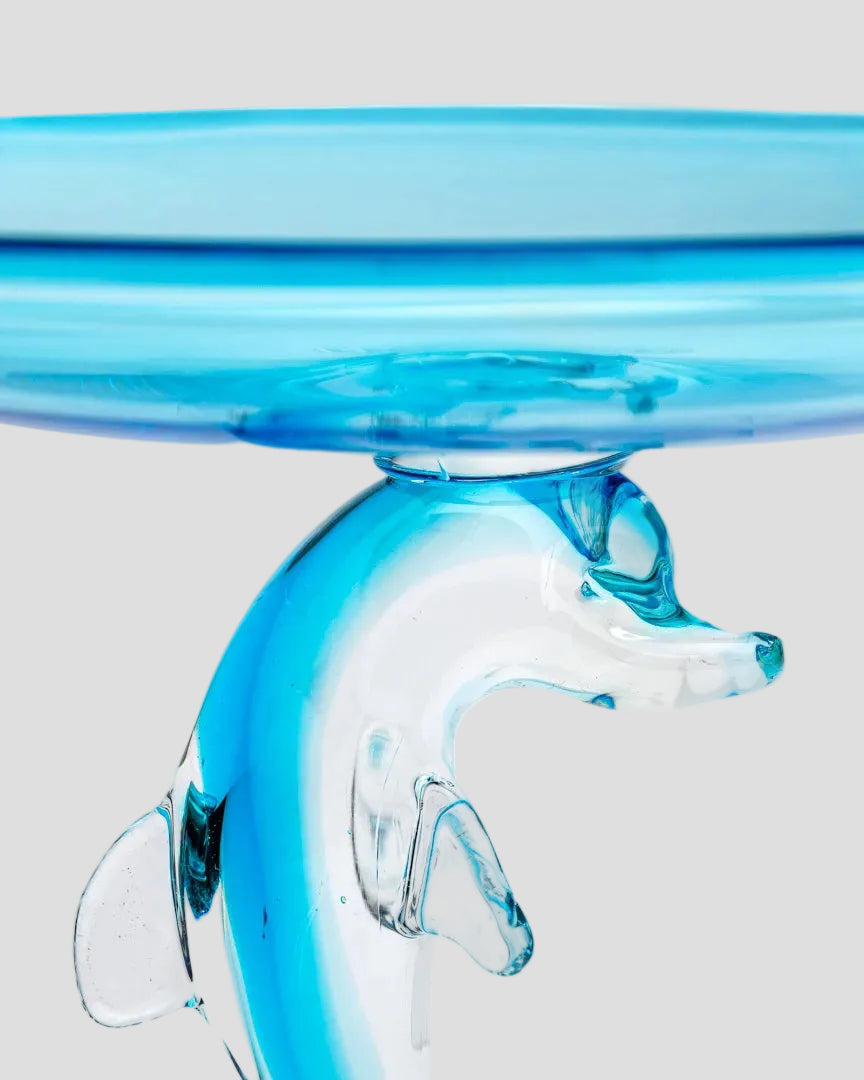 A close-up of a transparent blue glass object with a dolphin-shaped support and a curved blue rim, photographed against a white background.