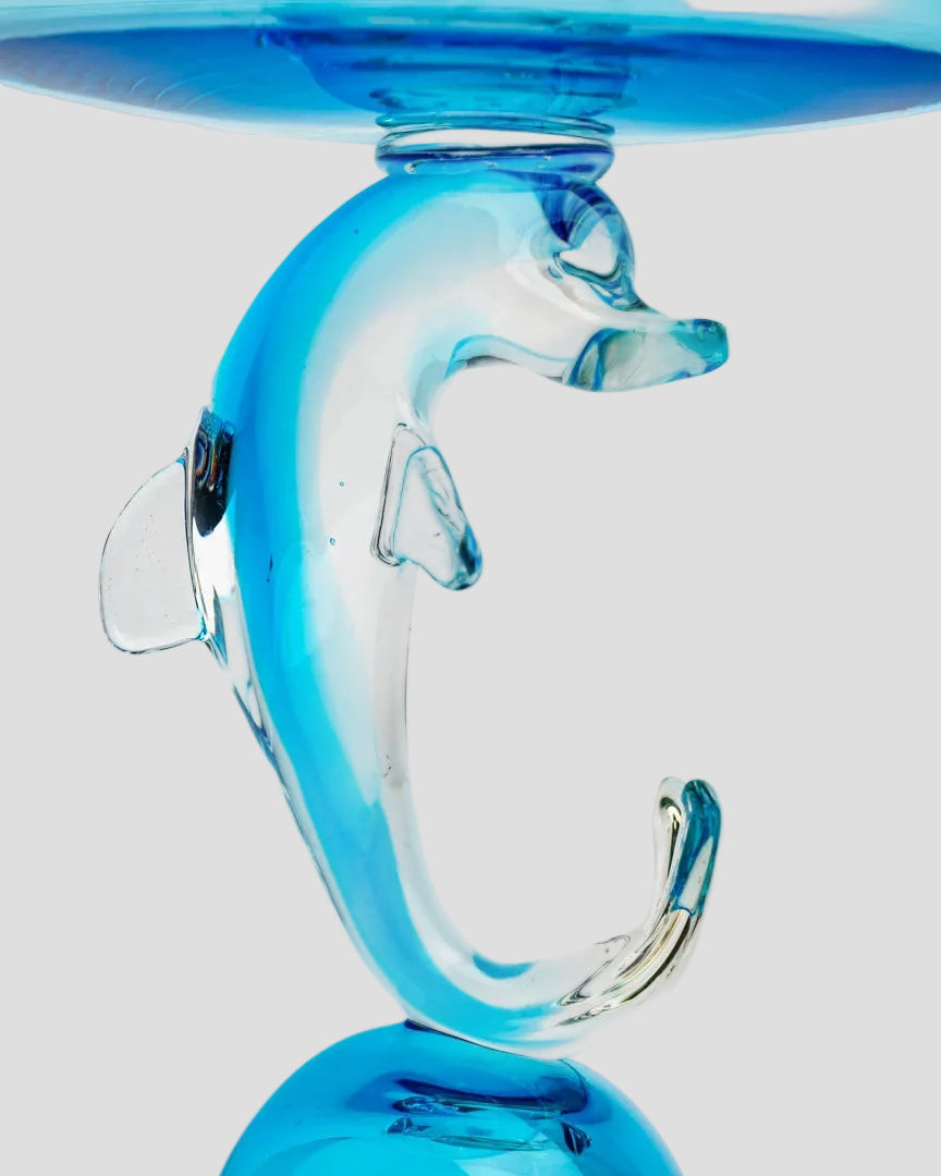 A close-up of a transparent blue glass dolphin-shaped stand or holder on a white background, with part of a matching blue glass rim visible at the top.