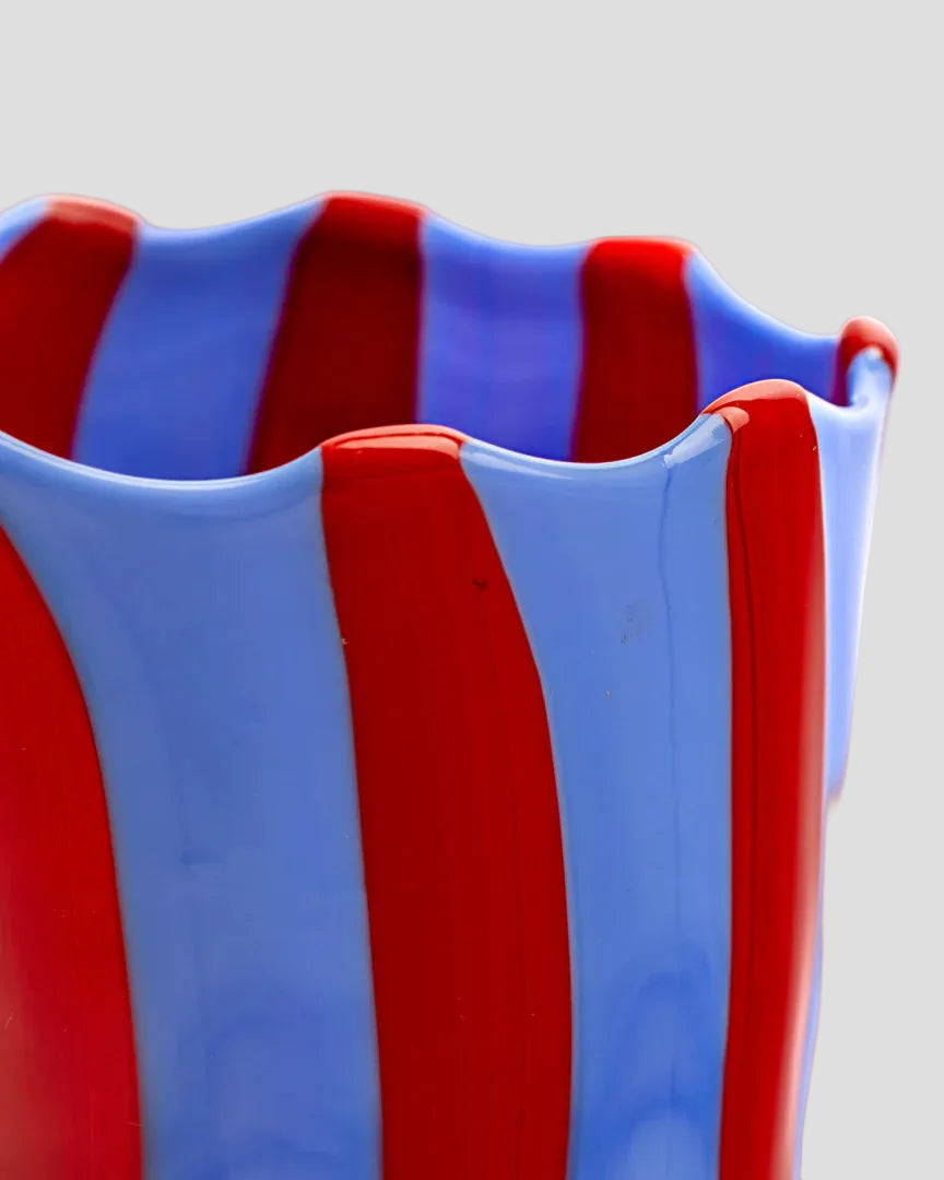 Close-up of a glass vase with a wavy rim, featuring alternating vertical stripes of glossy red and translucent light blue on a white background.