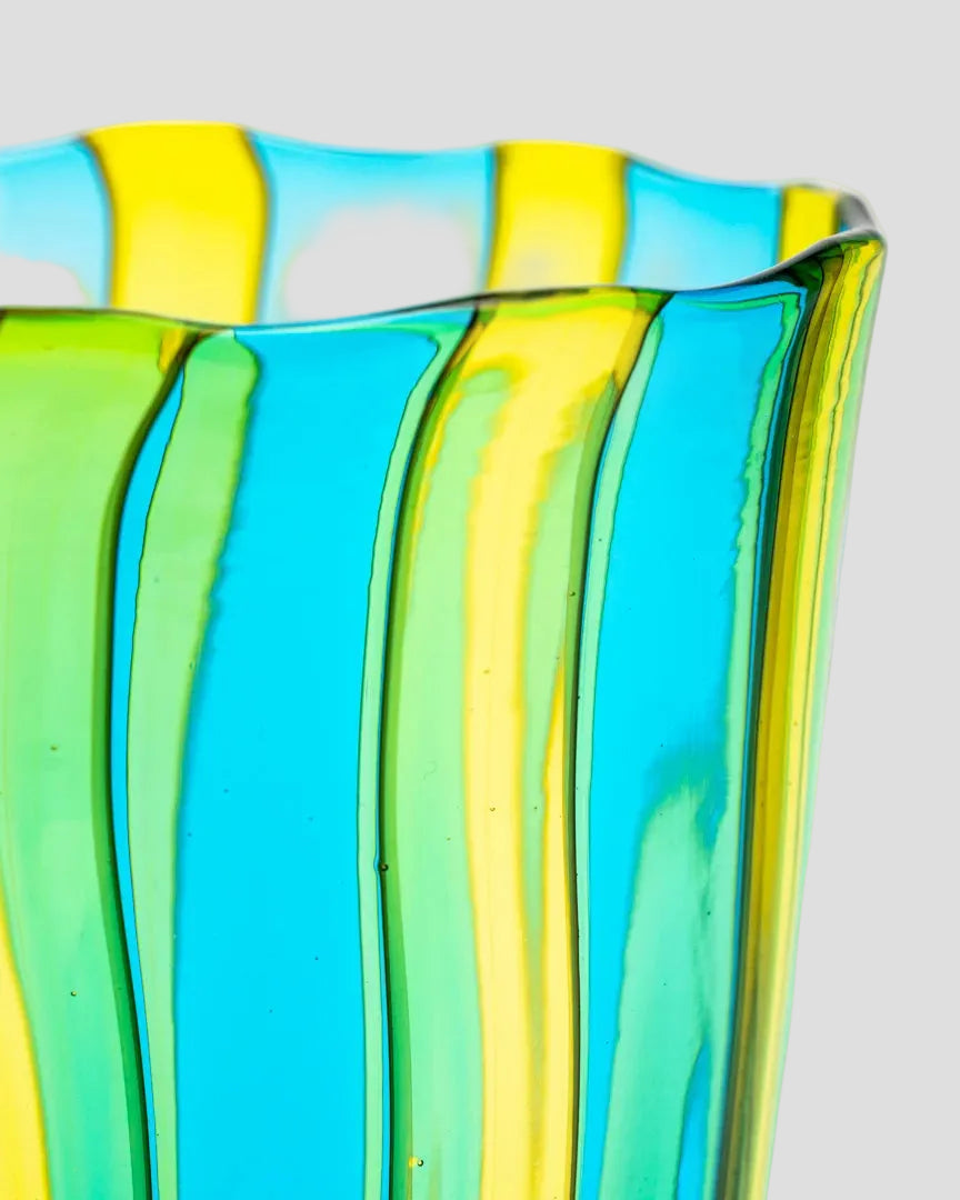 A close-up of a translucent glass vase with vertical fluted ridges and alternating bright blue and yellow-green glass bands, set against a plain white background.