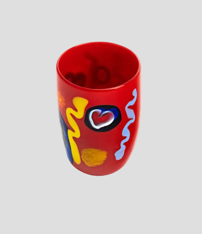 A red glass tumbler decorated with colorful abstract patterns including a heart, yellow and blue swirls, and a yellow circle, set against a plain white background.