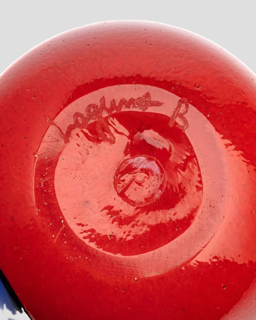 Close-up of the glossy red underside of a round glass vessel, showing a circular base with a hand-signed autograph and a small embossed logo.