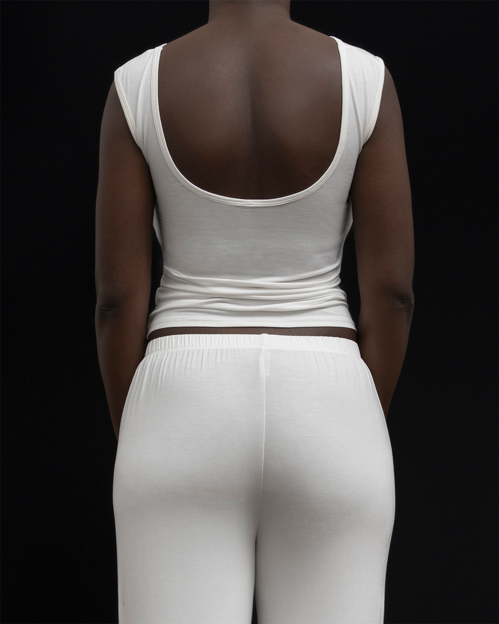 Back view of a model in Kye Recline Bateau Sleeveless, showcasing the open back design of the white bamboo boat neckline sleeveless tank, paired with white pants.